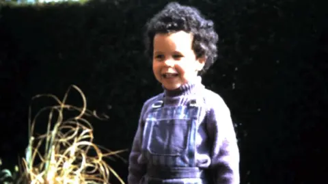 Vik Fielder Fielder as a young girl, with short black curly hair and wearing a purple jumper and blue, striped dungarees.