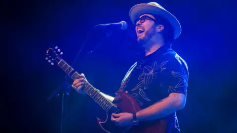 Getty Images A singer with the band the Cinelli Brothers in sunglasses and a hat plays his guitar in a patterned shirt in front of a microphone