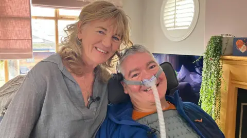 BBC The image shows a man and a woman positioned closely together indoors. The man is seated in a supportive chair, with a visible ventilation tube running from the lower part of the frame towards his chest. He is wearing a padded grey top with a blue outer layer. The woman is leaning in beside him and is wearing a textured long‑sleeved shirt with a microphone clipped to the front. Behind the two people is a round window with shutter-style blinds, and a fireplace with a wooden mantel.