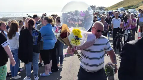 A crowd of people at a beach. A man with sunglasses holds a balloon and a bunch of flowers