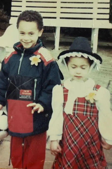 Chelsea Boyce A young girl is wearing a traditional Welsh dress and a daffodil pinny , and a hat, and is standing alongside a young boy who is wearing a coat and the same daffodil pinny.