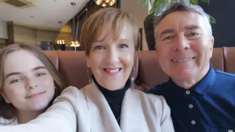 Family photo A selfie of Marianne with her husband and daughter taken in a restaurant. Marianne is wearing a cream coat over a black turtleneck jumper, with long silver earrings. Her husband is wearing a blue button up shirt and has short grey hair. Her daughter has longer light brown hair and pearl earrings. 