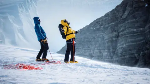 National Geographic/Freddie Claire Will Smith and Richard Parks are standing on a snowy mountain at a high altitude. Will Smith has raised his arms spread out and is looking up at the air.