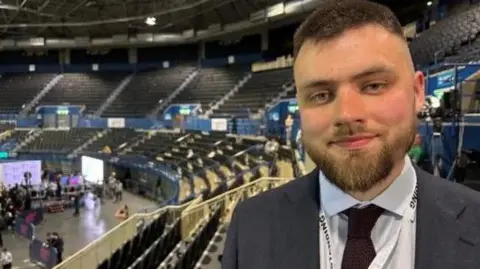 A young man smiles into the camera. He has brown cropped hair and a beard and moustache. He is wearing a grey suit jacket, white shirt and brown tie. A lanyard is around his necl. He is in an arena with lots of empty seats.
