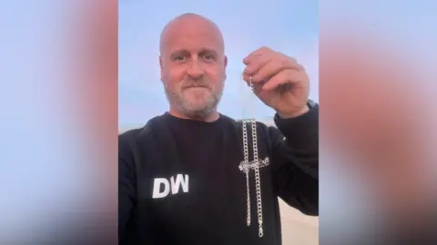 PA Media A man holding up a chain to the camera while stood on a beach.