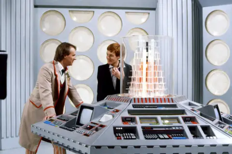 A still from the Doctor Who TV show showing two men standing on a futuristic set. On the left, a man wearing a long beige coat with red trim stands with his hands on a central console. He has light brown hair, short at the front and long at the back. He is talking to a second man who has ginger hair and is wearing a black suit. The console has six sides and is covered in keyboards, screens and switches, with a crystal-type stricture at the centre inside a glass box. The walls are grey with white, plate-shaped structures.