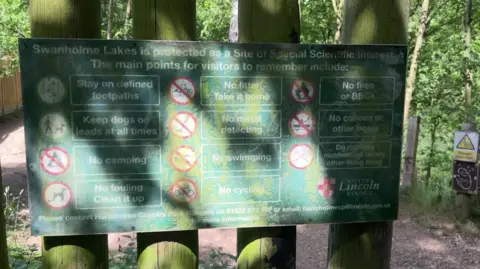 A close-up shot of a green board hung up on four wooden beams. It is in a nature park. it has 11 warnings on it.