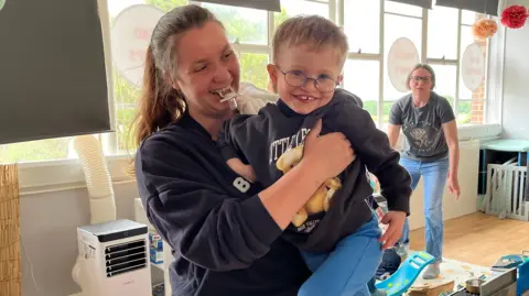 Charlotte holds Elijah on her hip and she has a dummy in her mouth. Elijah is about 4 years old wearing a dark blue sweatshirt and light blue trousers. He has glasses and is looking straight at the camera. Rachel has long brown hair pulled back in a pony tail. She has a dark blue sweatshirt on as well.