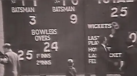 A screenshot from a video of a 1969 BBC film of Ireland playing against west Indies in cricket in which a number of people stand around an old style cricket scoreboard. The image is black and white