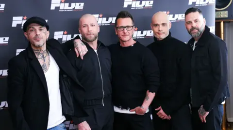 Members of the boyband Five, all in black, stand in front of a board with their group's logo