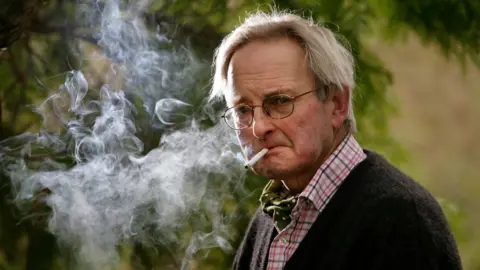 Allan Massie, in his late 70s, stands in front of a tree, smoking an un-tipped cigarette. There is a lot of smoke around him. he is wearing a dark brown cardigan, red and white checked shirt and green spotted cravat. He is staring at the camera.