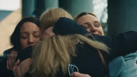 BBC/Alleycats TV Three women hugging. Two face the camera, with eyes closed. One faces away from the camera.