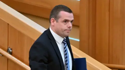 Douglas Ross walking out of the Holyrood chamber. He has short dark hair and is wearing a black suit, white shirt and blue and black striped tie