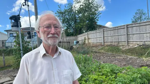 An elderly man stands in front of his vegetable garden with a wooden fence in the background.