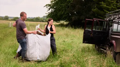 Andrew Montgomery Mr Allen (left) and Mrs Allen (right) are carrying a huge canvas bag full of sheep wool towards a vehicle.