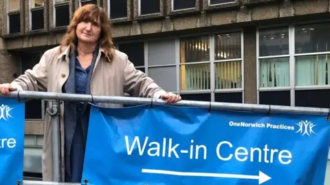 Lucy Galvin Lucy Galvin is wearing a beige overcoat on top of a blue dress. Her hands are resting on a metal railing, from which a blue "Walk-in Centre" sign hangs.