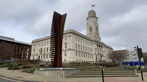 Barnsley town hall is a large, white stone building with a clock tower. Outside is steps and a large rust coloured sculpture which looks like a twisted hair comb