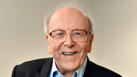 An elderly man wearing glasses and smiling.