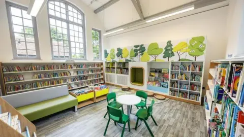 A room with bookshelves on all the walls and a small circular table in the middle, with a large window on the left-hand side