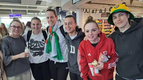 Six of Wales' women football team stand with their arms around eachother, smiling towards the camera in the arrivals lounge in Bristol airport after landing home from Dublin on Wednesday. Some wear Wales FC hats, hoodies and scarfs while Laura O'Sullivan wears a Welsh flag tied around her neck.