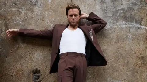 Blackpool Council Olly Murs is wearing a brown suit and white T-shirt. He has brown hair and a brown beard.