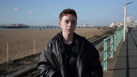 Lysander Pierce wears a black leather jacket and stands on Brighton seafront with the West Pier and the shingle beach in the background.