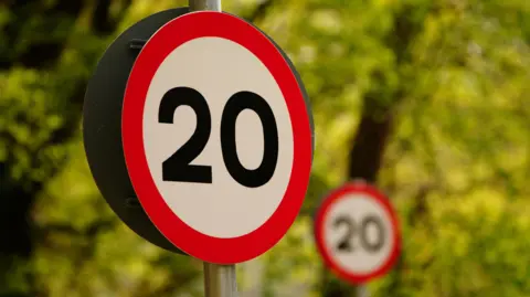 A big 20mph road sign with another in the distance, with trees in the background