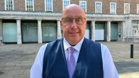 A bald man wearing glasses and a waistcoat, purple tie and white shirt. He is stood in front of the Nottinghamshire County Council HQ