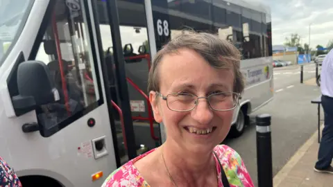 John Devine/BBC Michelle Rham has short brown hair and gold framed thin glasses on, she is smiling and wearing a pink floral top with a gold necklace around her neck, behind her is the mini bus, white in colour.