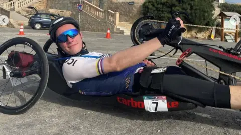 Dr Gordon is lying down in a hand cycle. He is wearing cycling glasses, helmet and clothing.