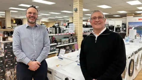 Stephen Hill and Mark Raisey are standing in front of washing machines in the shop, looking at the camera. They are both smiling. 