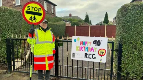 Reg Bown Reg dressed in high-vis uniform, holding his STOP sign. He is smiling, standing next to a sign which reads 'Happy 90th Birthday Reg'.