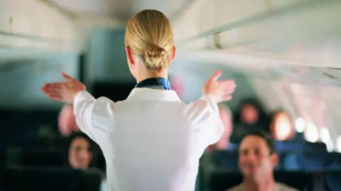 An air stewardess facing away from the camera shows passengers on a jet the emergency exist during a safety briefing. She is wearing a white shirt and her blonde hair is in a tight bun