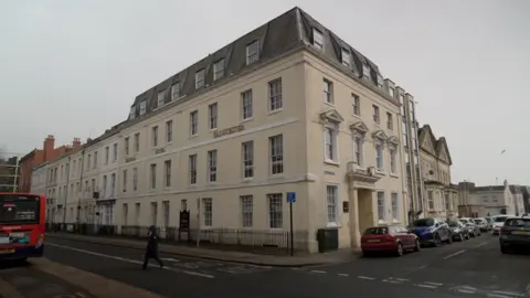 Two sides of a lightly coloured three storey building in the Georgian style with cars parked in front and a passing bus.