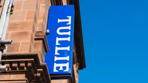 One of the blue signs with the word Tullie in white that has now been removed. It is attached high up the side of a red stone building.