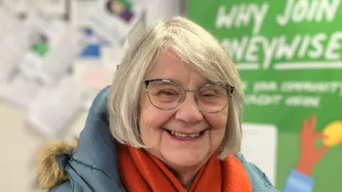 Christine Shearer has grey medium length hair and round black glasses. She is smiling and has an orange scarf and a blue jacket. 