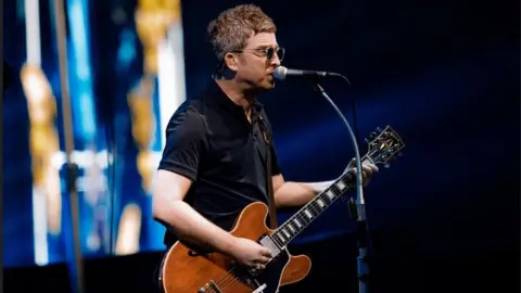 Big Brother Recordings Noel Gallagher wears sunglasses as she plays his Gibson guitar and sings on stage. He is wearing a dark coloured short sleeved top.