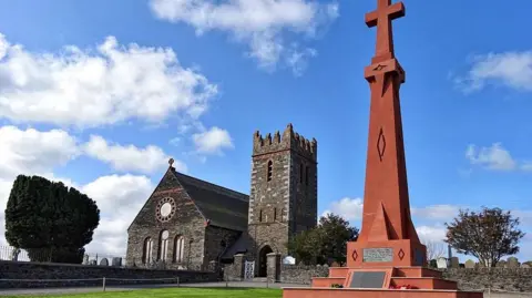 Andreas War Memorial - a red sandstone plinth with a cross on top - stands in front of Andreas Church and church tower, which are bult of dark grey stone.