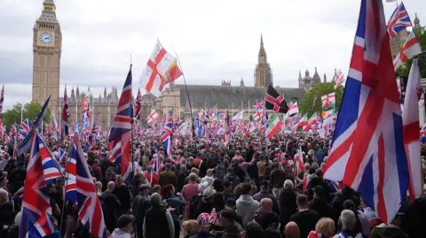 PA Media People take part in the Unite the Kingdom march in central London. A large crowd holding a range of banners and flags aloft is pictured in front of Big Ben. Mostly visible are Union flags.