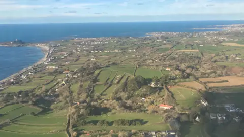 BBC An aerial view of Guernsey - fields, trees and houses with the sea and blue skies surrounding it.