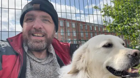 Open University David Scott with speckled grey and brown beard, black beanie hat, grey jumper and red and grey coat alongside a golden labrador