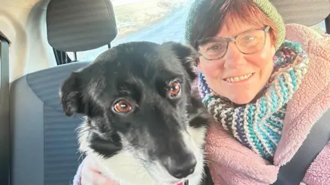 Susan Taylor A woman with red hair in a green hat with a colourful scarf and coat has her arm around a black and white dog with brown eyes. They are sitting on the front seats of a car and the woman has a seatbelt on
