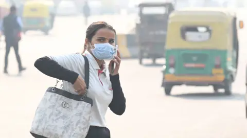 A person holds a phone to their ear as they walk down a street in Noida while wearing a face mask with tuk tuks in the background driving down the street, taken in November 2025.