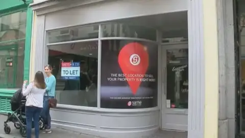 A picture of a grey shop front with a to let sign. Standing next to it are two women in conversation with a pram.