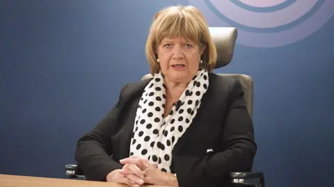 PA Baroness Heather Hallett sits in the inquiry chair wearing a black suit and spotty scarf, with the UK Covid-19 Inquiry logo on the wall behind her