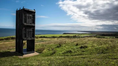 BBC The Pit Cage Monument stands on a grassy field with the coast visible in the distance. It is a sunny day with a clear sky over the sea and clouds towards land.