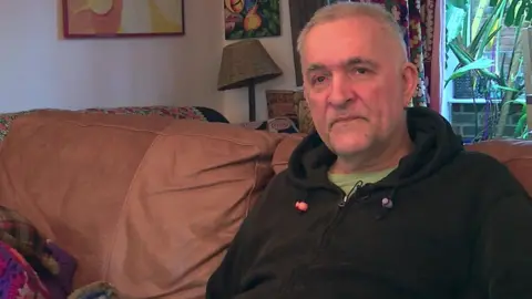 A man sat on a sofa. He is wearing a black hooded top and is looking directly at the camera. 