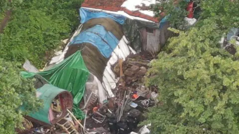 CPS A police drone photo shows a ramshackle scene of various buildings in a wooded area. One is made of green, brown and white tarpaulins. Another is a wooden barrel with wooden steps to an open door. another is a shack building with a red tile roof. There is rubbish strewn all around the property