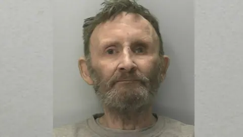 Devon and Cornwall Police A mugshot of Mark Fitzgerald. He has a grey beard and grey hair. He is stood in front of a grey wall.