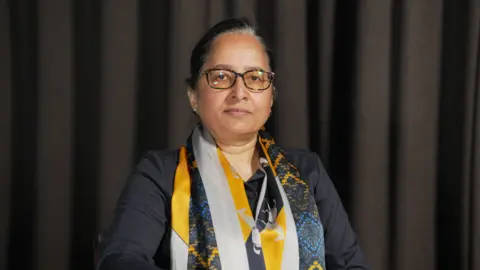 This image shows a woman with black hair and brown eyes, she's wearing black glasses and is stood in front of a dark grey curtain. She's wearing a black top and a patterned yellow and blue scarf. 